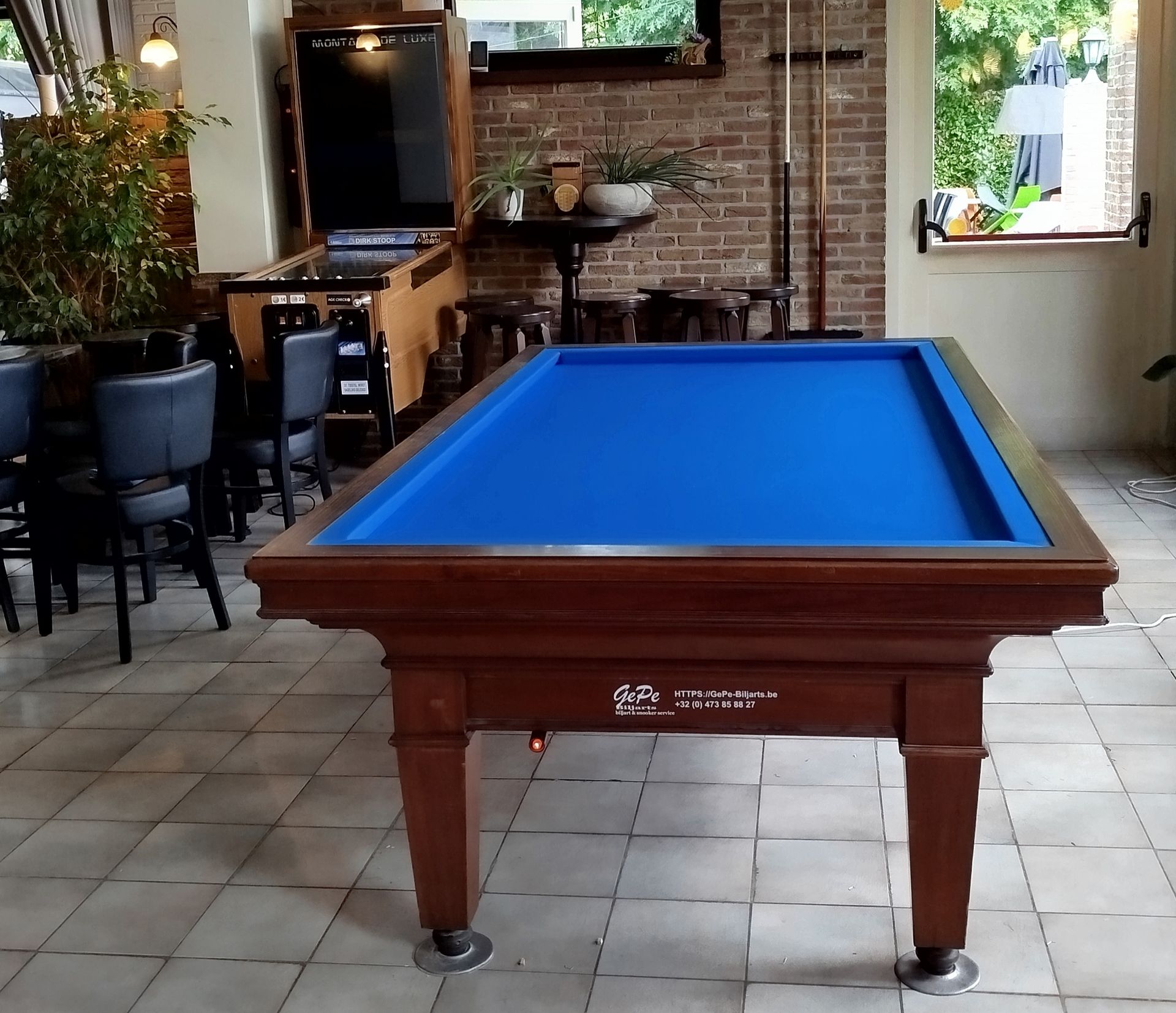 Mavi çuhalı ve koyu ahşap kasalı restore edilmiş bilardo masası, bar tabureleri ve fayans zemini olan bir kafede.